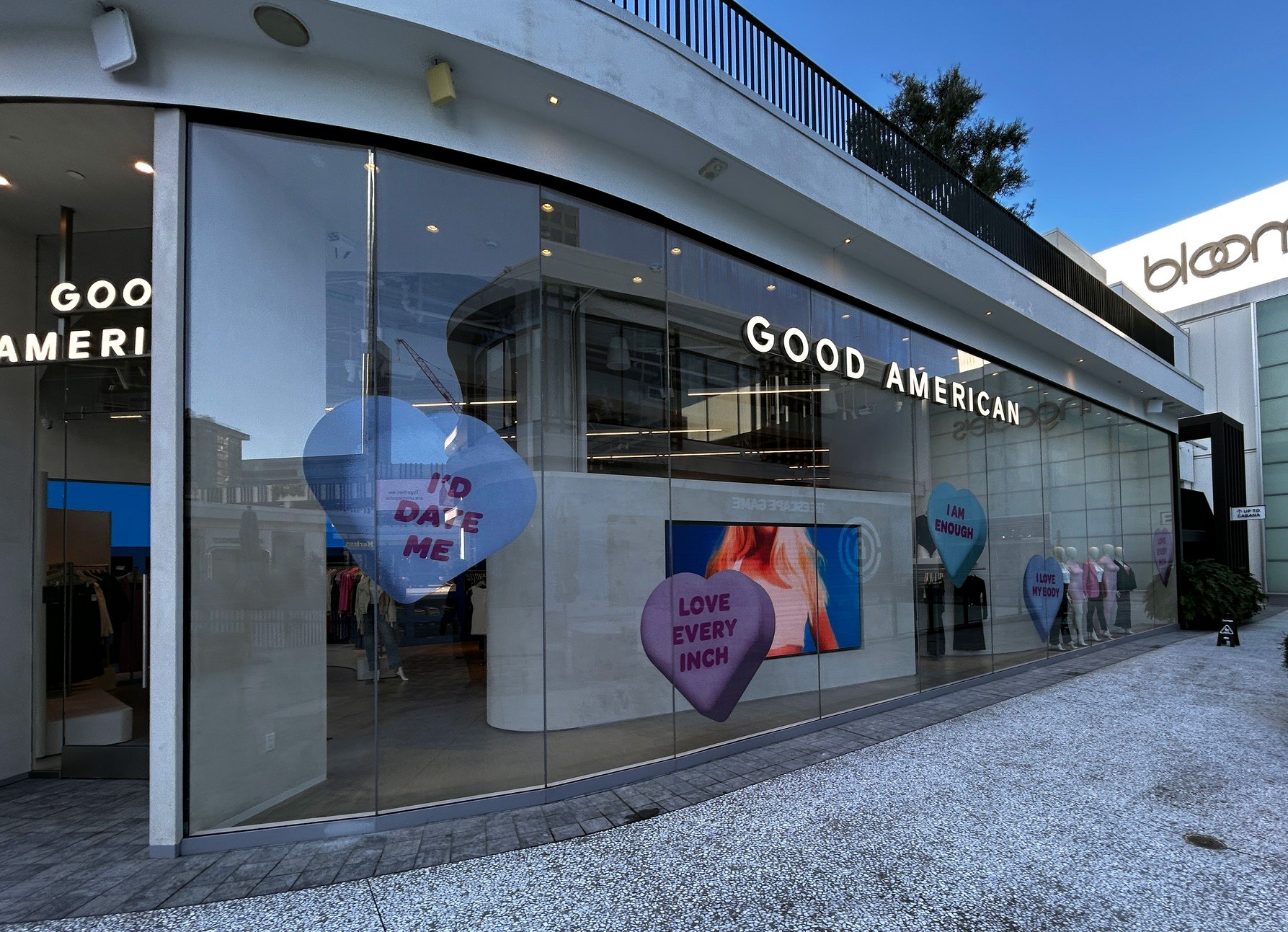 Good American retail store exterior at Century City mall with large heart-shaped window decals reading 'Love Every Inch,' 'I'd Date Me,' and 'I Am Enough'
