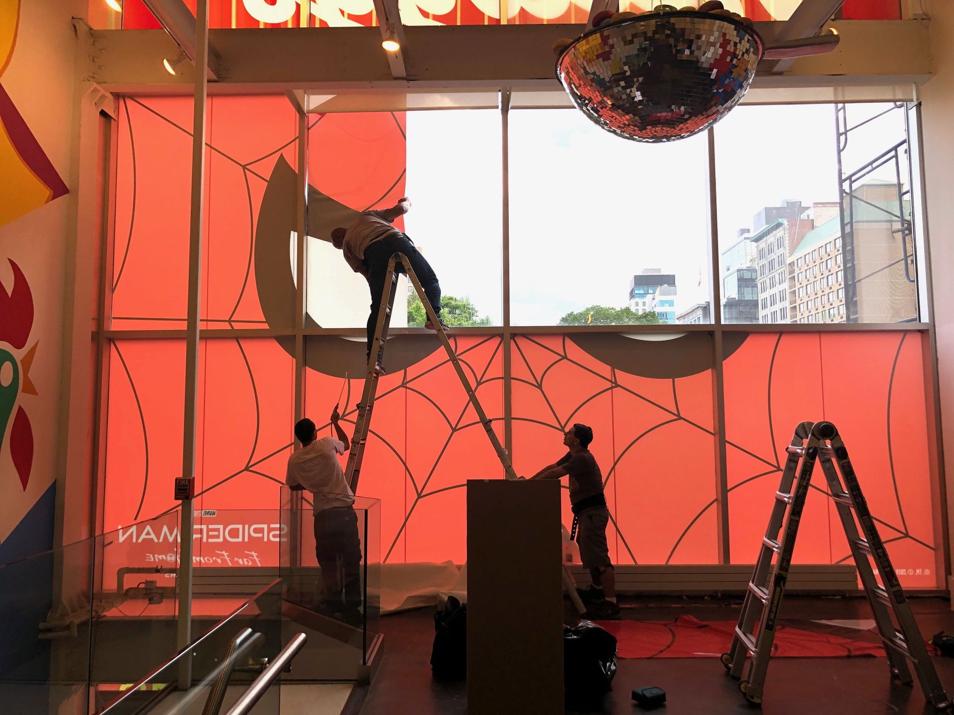 Installation crew applying a large Spider-Man Far From Home window graphic with spiderweb design inside a retail store, workers on ladders during installation