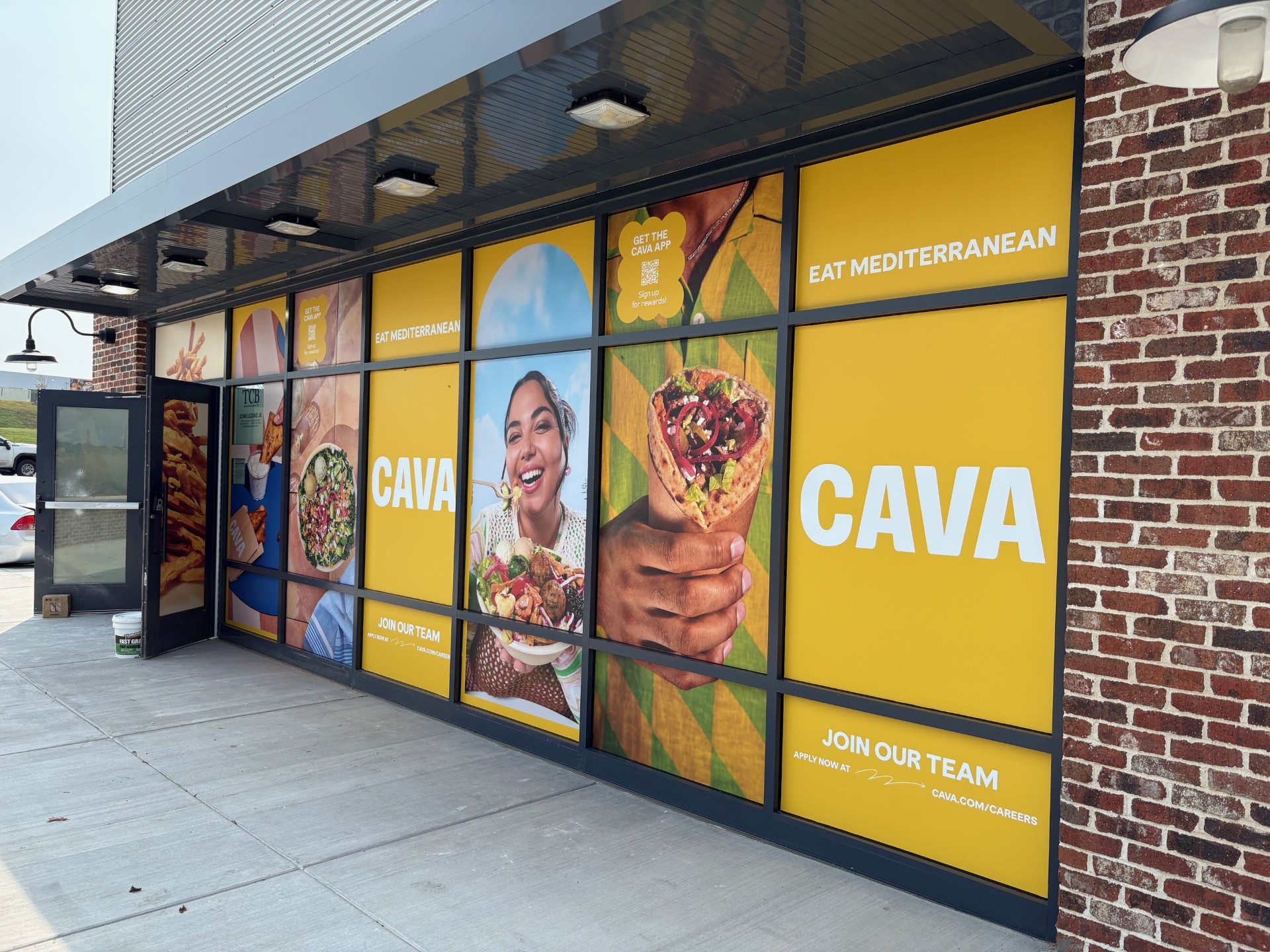 CAVA restaurant exterior window graphic installation in Nashville, Tennessee featuring bold yellow branded panels with food photography, 'Eat Mediterranean' messaging, and a 'Join Our Team' recruiting panel