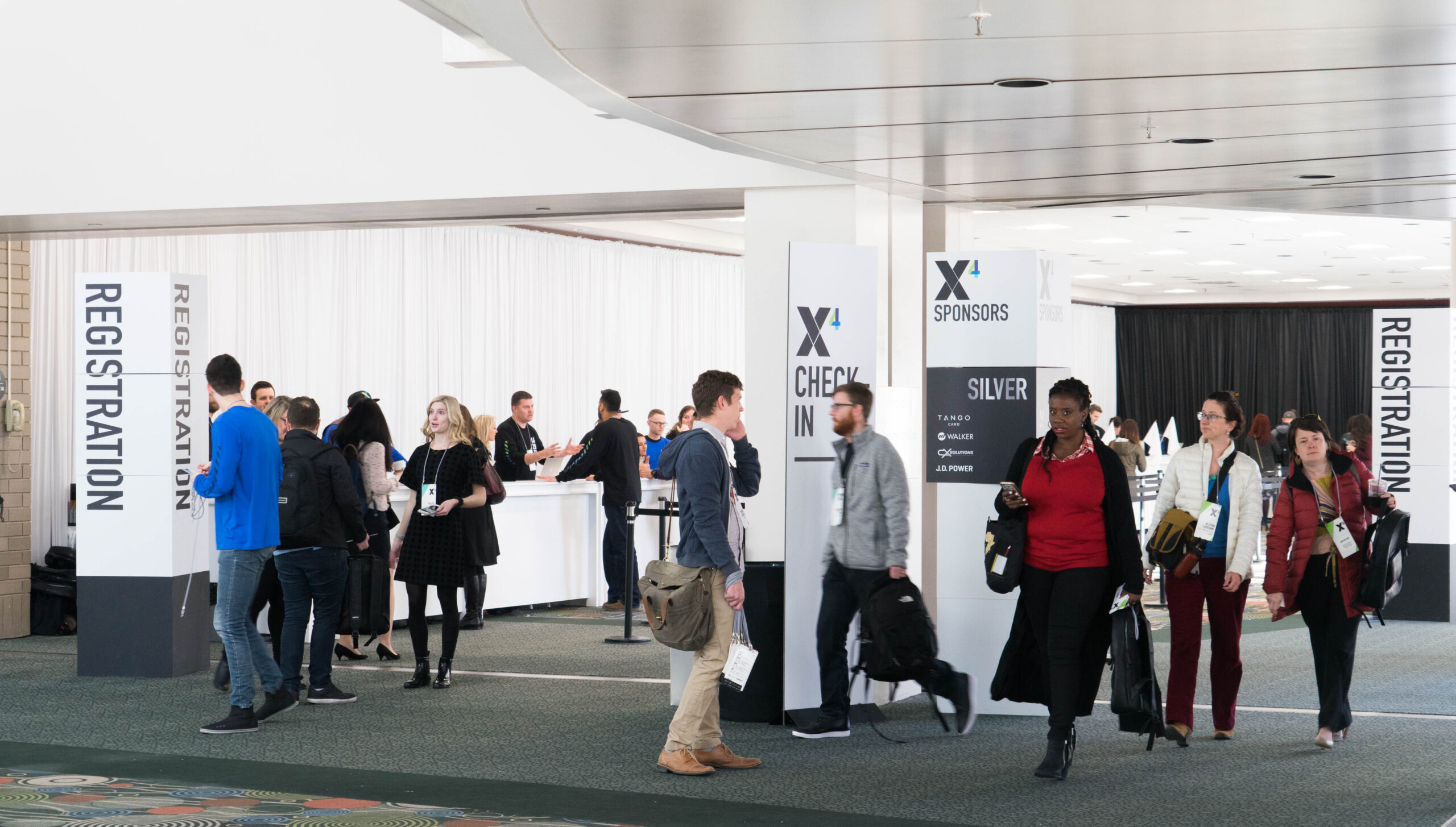 Busy conference registration area with attendees checking in at branded X4 event, featuring large vertical registration banners, a sponsors display, and a Silver sponsors board