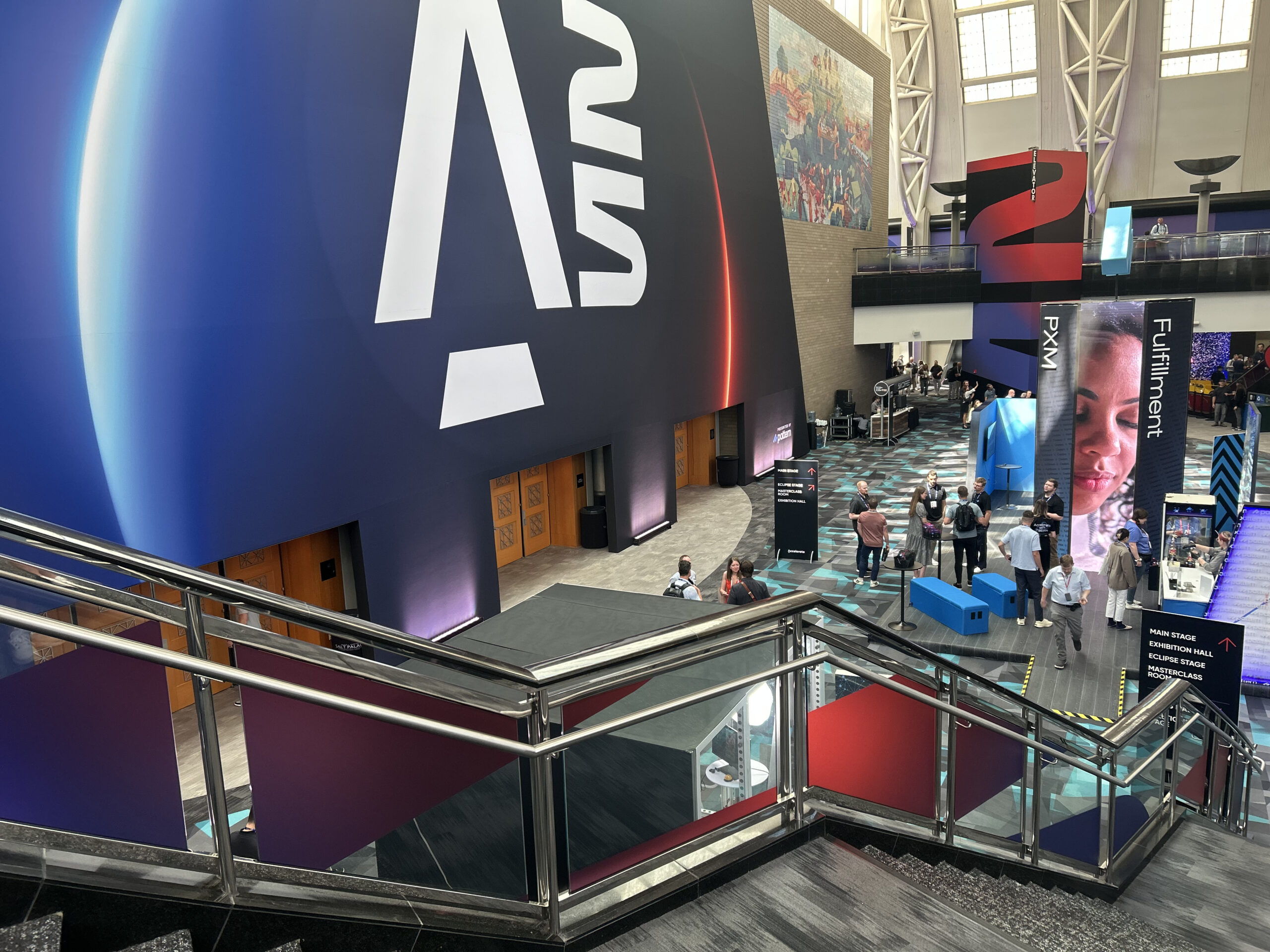 Large convention center event space with oversized branded signage, illuminated displays, and attendees on multiple levels, featuring PXM and Fulfillment themed exhibit areas and a Main Stage directional sign