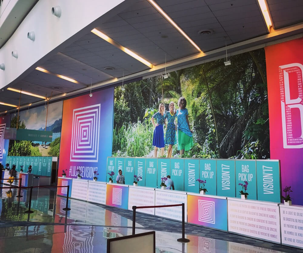 LuLaRoe Vision '17 conference event space featuring large-format tropical backdrop wall graphic, colorful geometric step-and-repeat banners, bag pickup signage, and attendees in a convention center lobb