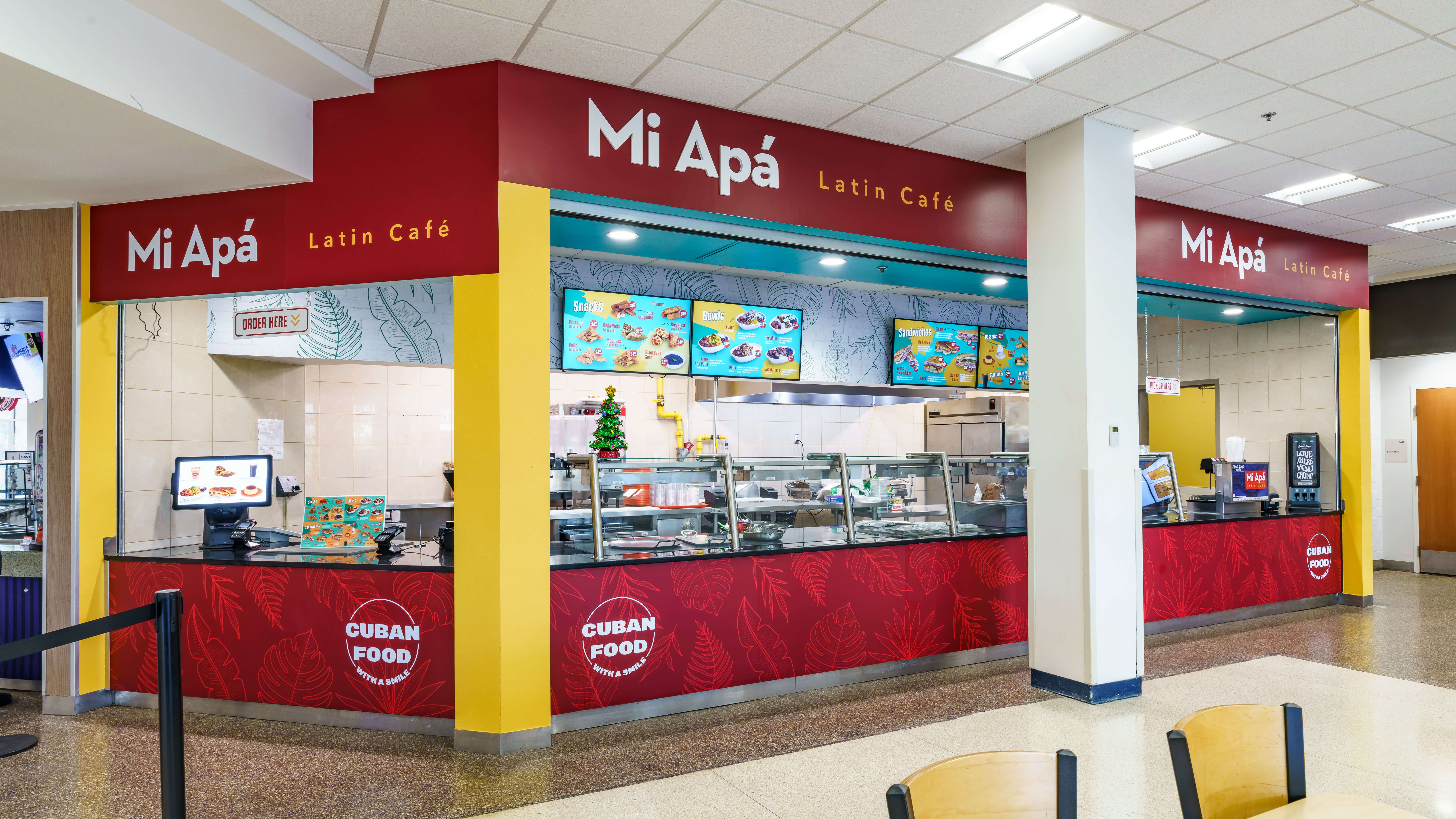 Mi Apá Latin Café branded food service counter at the University of Florida featuring bold red and yellow counter wraps, Cuban Food medallion signage, overhead branding arch, and digital menu boards