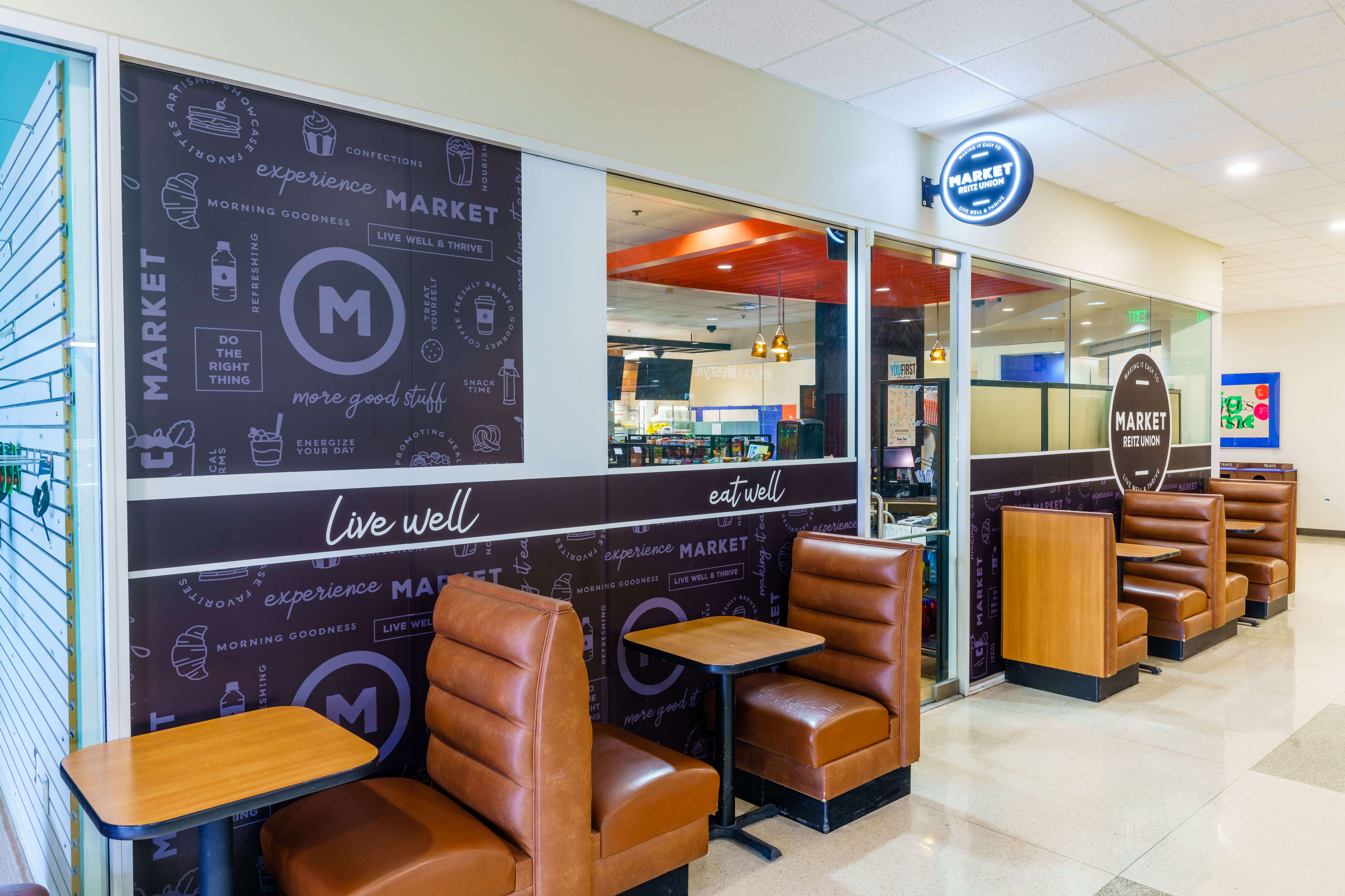 University of Florida Market Rest Union branded interior wall graphics featuring dark wall wrap with 'live well, eat well, experience Market' messaging, large M logo, and illustrated food and beverage icons, with leather booth seating in the foreground