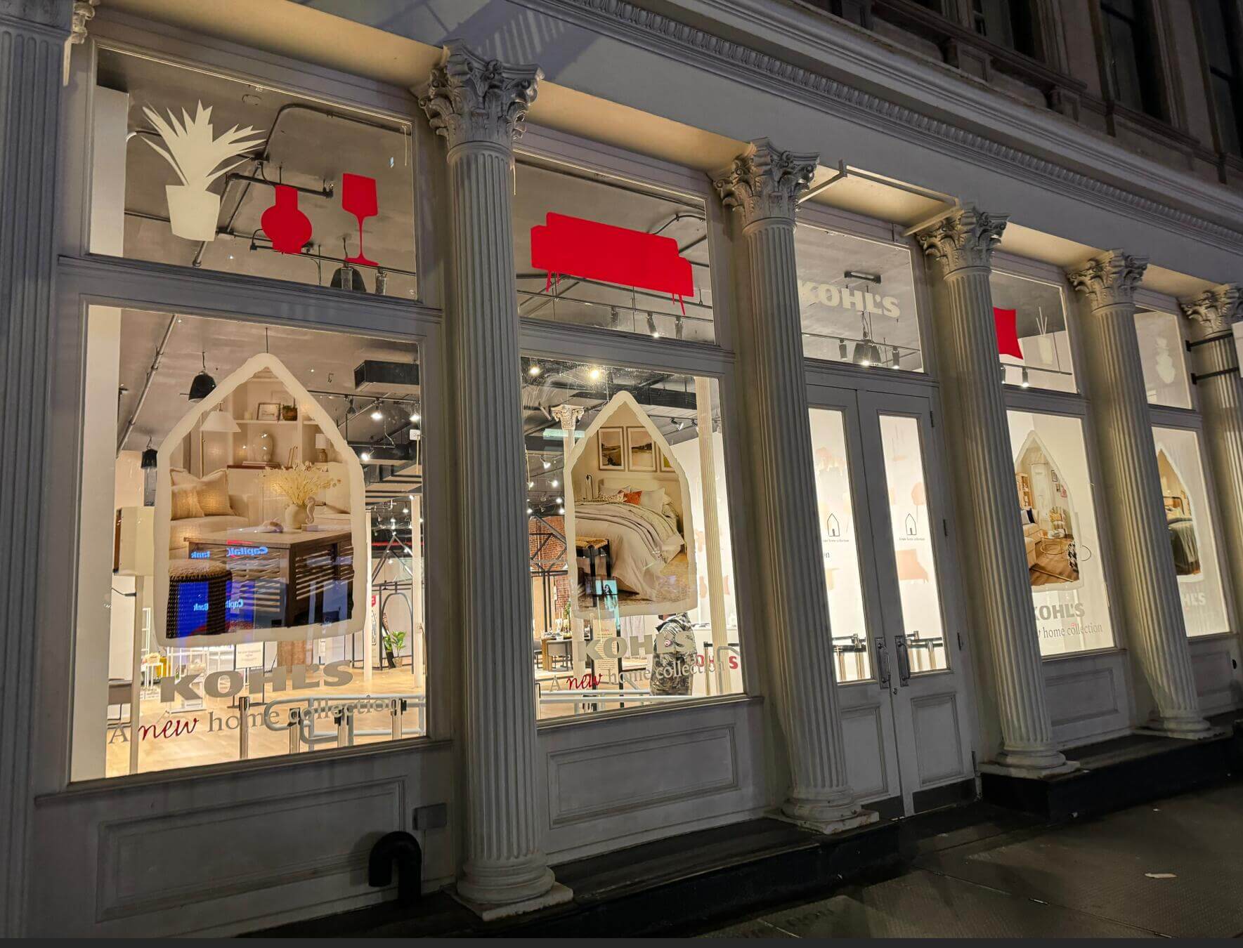 Kohl's 'A New Home Collection' storefront window display on a classic architectural building facade, featuring bold red graphic panels, arched window displays with home décor vignettes, and Kohl's branding across multiple street-level windows