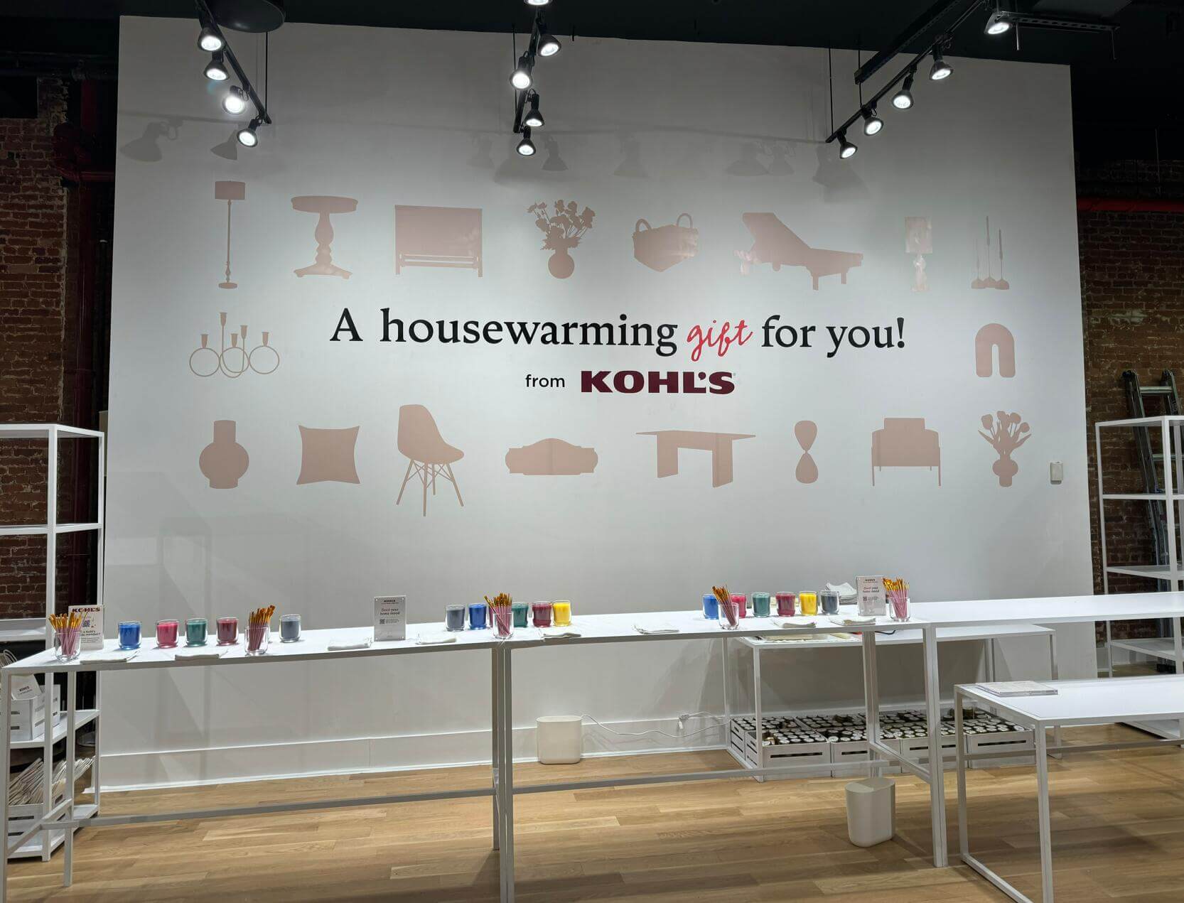 Kohl's 'A housewarming gift for you!' large backdrop in a styled retail setting, surrounded by illustrated home furnishing silhouettes and colorful gift items on a table in the foreground