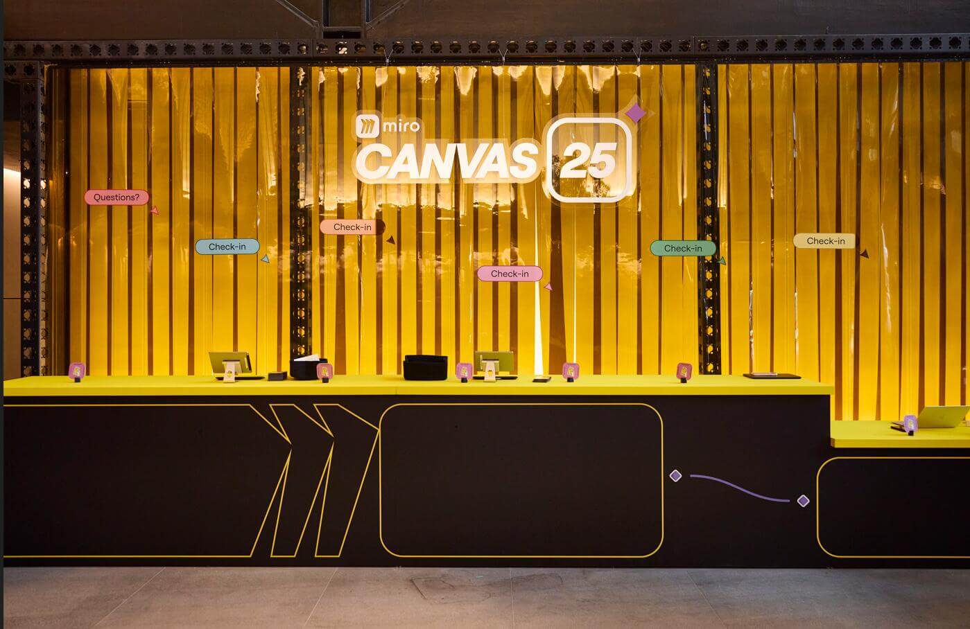 Miro Canvas 25 event check-in counter viewed straight-on, featuring illuminated Canvas 25 signage, gold tinsel curtain backdrop, and multiple staffed check-in stations with yellow counter detailing