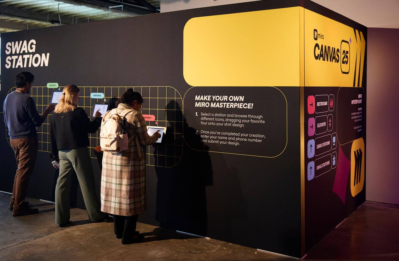 Attendees interacting with a Miro Canvas 25 branded Swag Station activation booth, featuring a 'Make Your Own Miro Masterpiece' interactive shirt design experience with touchscreen kiosks