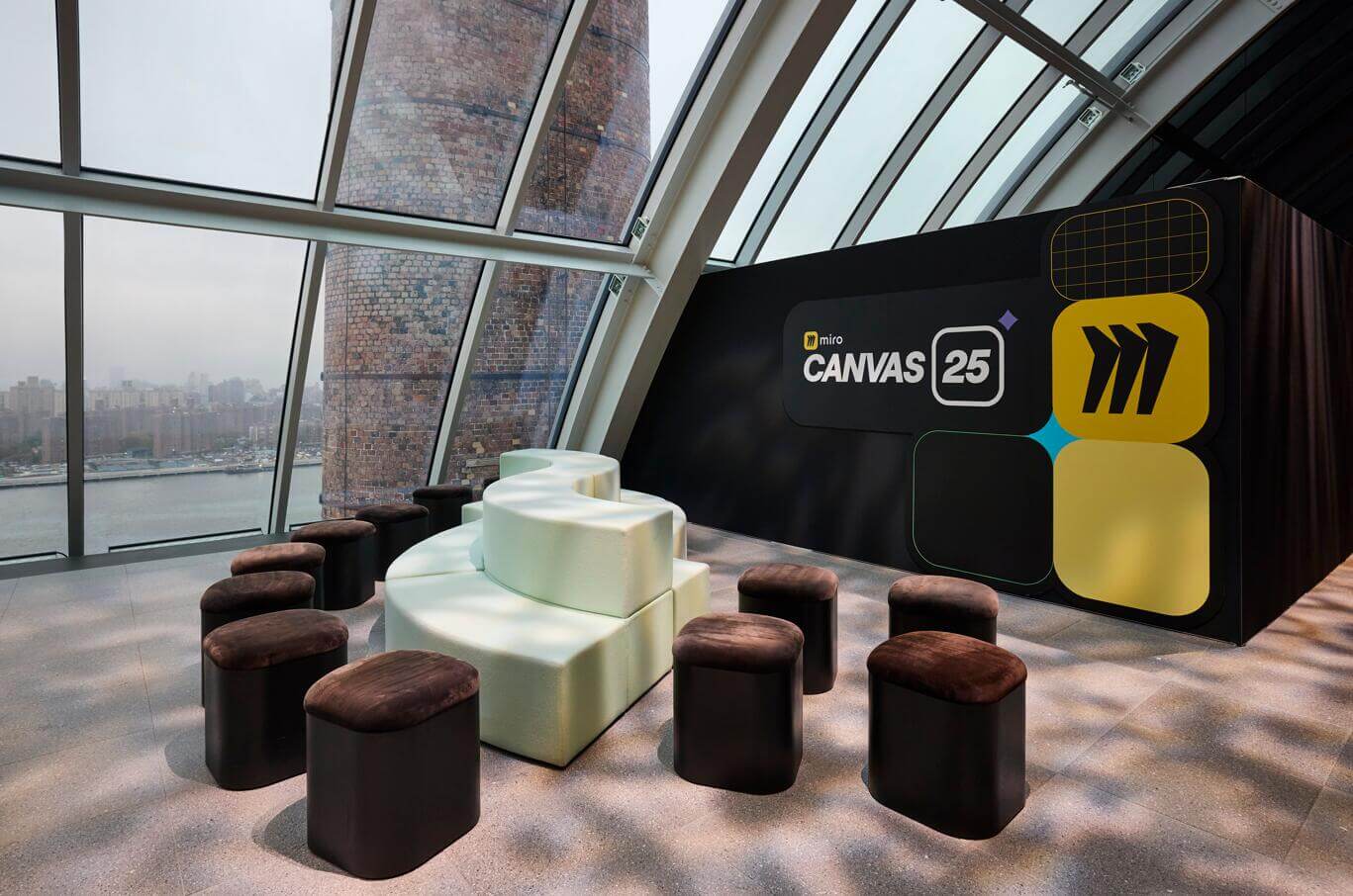 Miro Canvas 25 branded event lounge area with curved white modular seating, dark ottomans, a large Canvas 25 backdrop display, and floor-to-ceiling windows overlooking a city skyline