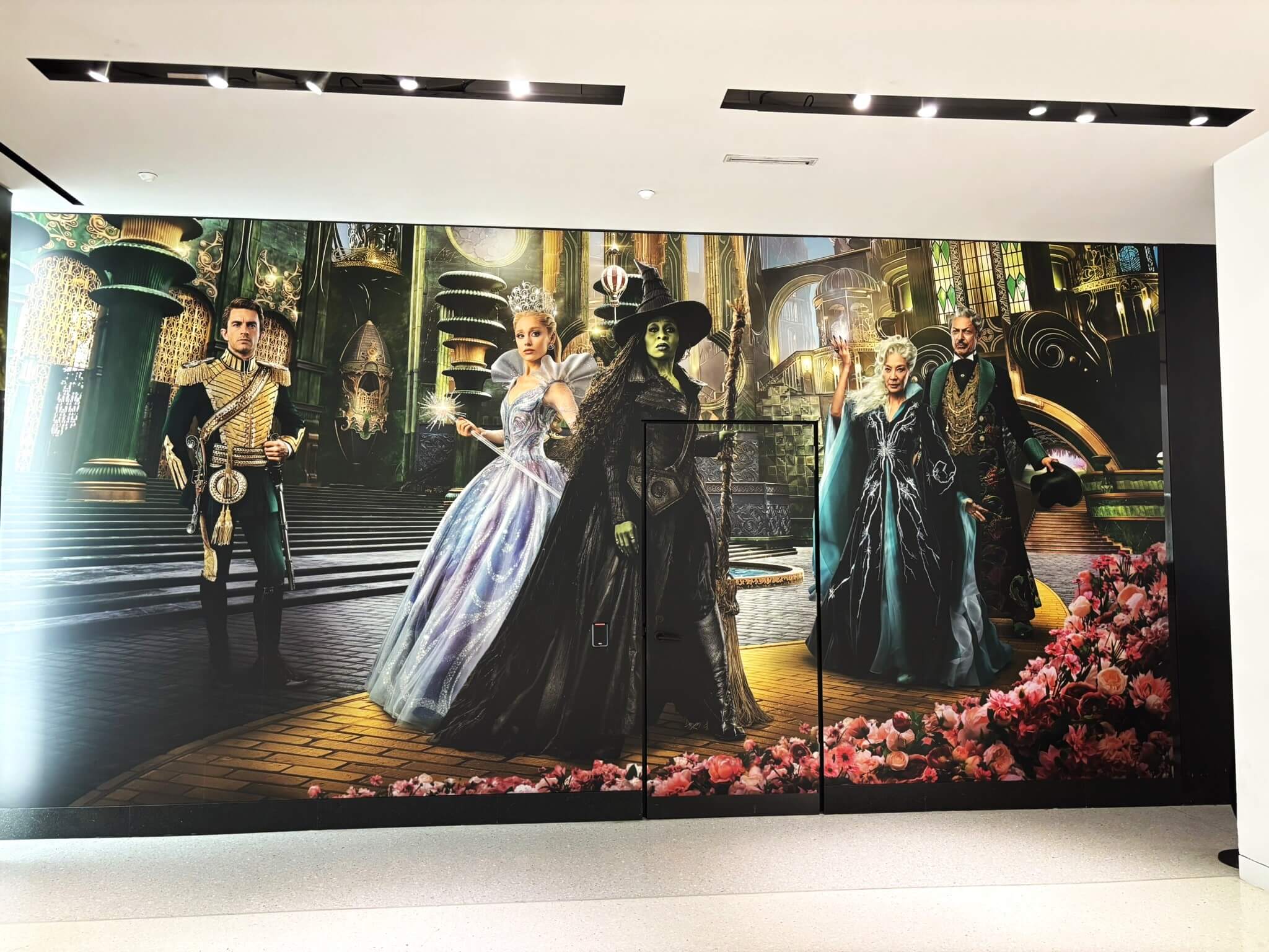 Here's what I'd suggest for the Wicked vinyl wall graphic: Alternative Text "Large-scale Wicked movie promotional vinyl wall graphic installation showing the full cast in an ornate fantasy setting, displayed across multiple panels in a retail or theater environment